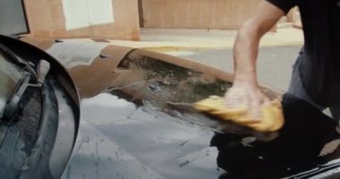 Kapalı tişörtlü zayıf adam siyah arabayı paçavra ve bol suyla yıkıyor.