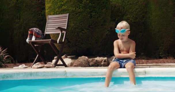 Мальчик сидит перед бассейном и ныряет в голубую воду — стоковое видео