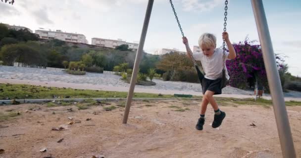Leuke baby blonde jongen swinging op ketting swing in speeltuin in bloeiende park — Stockvideo
