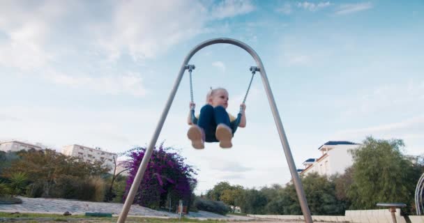 Slow motion cute european baby girl swinging on swing on chains in city park — Stock Video