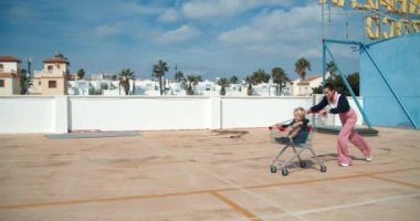 Genç mutlu anne alışveriş arabasıyla koşuyor ve çocuk park yerinde oturuyor.