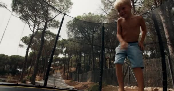 Little happy boy is jumping on trampoline installed in forest of rural terrain — Stock Video