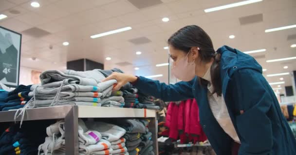 Mujer joven con máscara de protección facial buscando el tamaño de la ropa en la tienda — Vídeo de stock