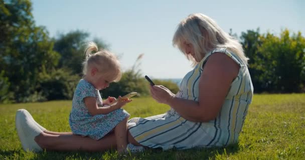 Babcia z wnuczką córeczką siedzi w trawie i używa telefonu komórkowego — Wideo stockowe