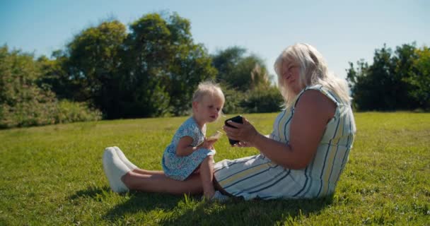 Abuela con la nieta de la niña sentada en la hierba y el uso de teléfono móvil — Vídeos de Stock