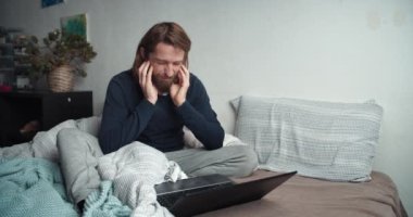 Kendini kötü hisseden serbest çalışan bir adam kanepeye oturmuş öksürüyor ve bilgisayarla çalışıyor.