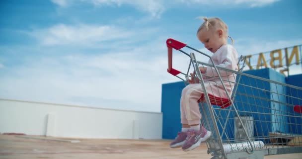 Χαριτωμένο κοριτσάκι κάθεται και χαμογελά σε σούπερ μάρκετ καλάθι αγορών σε εξωτερικούς χώρους — Αρχείο Βίντεο