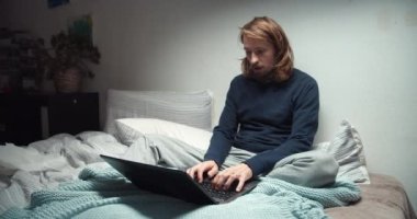 Kendini kötü hisseden serbest çalışan bir adam kanepeye oturmuş öksürüyor ve bilgisayarla çalışıyor.