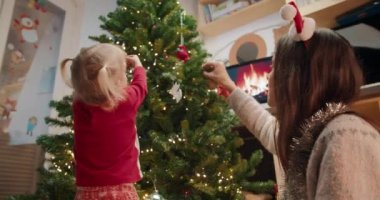 Çocuklu genç anne Noel ağacını akşam oturma odasında süslüyor.