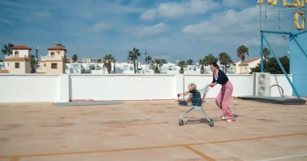 Junge glückliche Mutter läuft mit Einkaufswagen und Kind auf Parkplatz — Stockvideo
