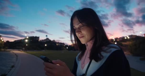 Улыбающаяся молодая привлекательная женщина пользуется смартфоном в парке на закате — стоковое видео
