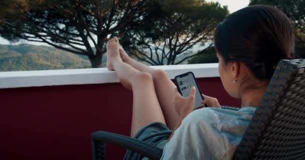 Amplio tiro de mujer joven en el balcón al atardecer utilizando el teléfono inteligente charlando y sonriendo — Vídeos de Stock