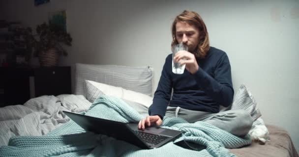 Niedobrze czuje się młody wolny strzelec siedzi w łóżku i pije wodę za pomocą laptopa — Wideo stockowe