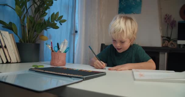 Счастливый улыбающийся мальчик рисует на бумажном листе цветным карандашом вечером — стоковое видео