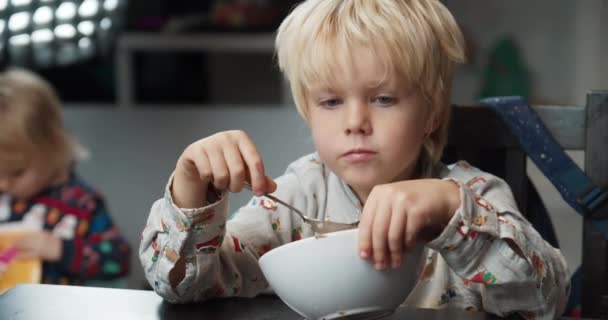 Child boy eats porridge using spoon in dinning room after school and speaking — Stock Video