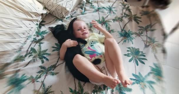Весела дівчинка лежить на ліжку з чорною домашньою кішкою і сміється — стокове відео