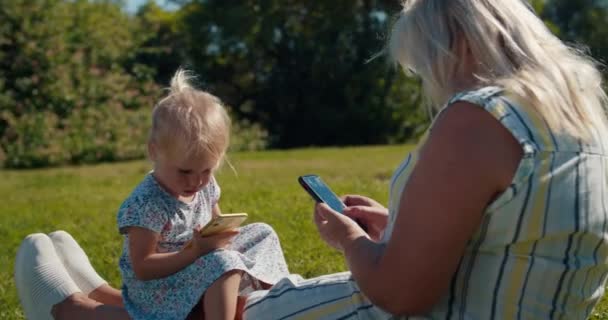 Büyük anne ve küçük kız torun çimenlerde oturuyor ve cep telefonu kullanıyor. — Stok video