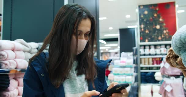 Young beautiful woman using phone during shopping in store and wearing face mask — Vídeo de stock