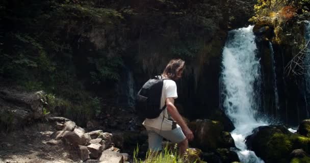 滝へのバックパックを歩くと彼の手を上げるとインスピレーションを男性旅行者 山のハイキングでアクティブなライフスタイル インスピレーションを受けた屈託のない男は自然風景を楽しむ 山のきれいな水の川を流れる — ストック動画