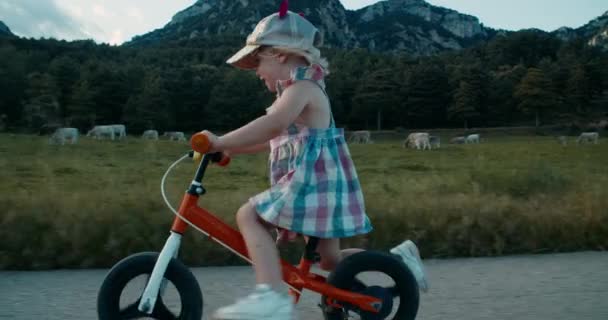 村の夜にケアフリー幸せな幼児の女の子がバランス自転車に乗る 牧草地に沿って山の景色を望む空の田舎道で自転車をサイクリング子供 田舎での積極的な生活 — ストック動画