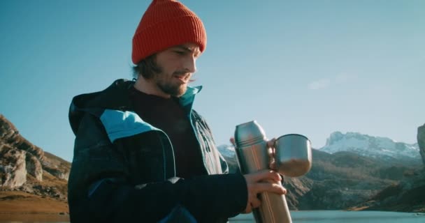 壮大な山の景色で魔法瓶から熱いお茶やコーヒーを注ぐ男性旅行者 ハイカーは春の旅で休憩します 男はバックグラウンドで雪のサミットで休暇中の道路の旅を楽しむ — ストック動画