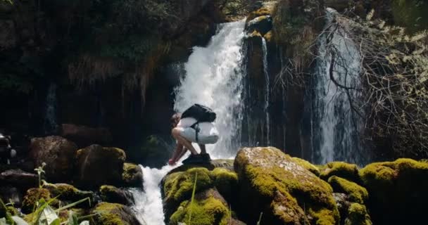 滝の近くでハイキングの冒険をしている男性旅行者 純粋な山の川に顔を洗ってリュックを持つ若い大人の男 夏の旅で渓流から水を集めるハイカー — ストック動画