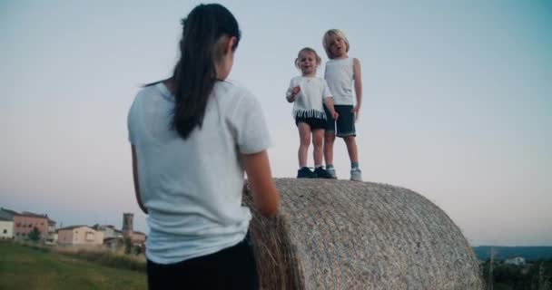 田舎の夏休みに一緒に幸せな家族 母親の撮影子供干し草のロールやベールの上に立つ 夕暮れ時の農村環境で子供の写真を撮るためにスマートフォンを使用して若い大人の女性 — ストック動画