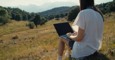 Wide shot young adult woman work using laptop on hike journey sitting on mountain. Freelance creative worker type on notebook keyboard with epic landscape view. Girl chatting with friend on travel