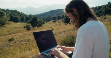 Medium shot of male freelance worker typing text message or notes on computer keyboard. Traveler on summertime journey using technology. Guy sitting on beautiful landscape with notebook.