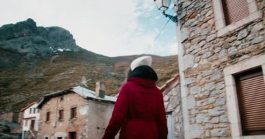 Female traveler exploring mountain village on vacation travel in red puff jacket. Woman walk on street of remote settlement in natural park on journey adventure. 