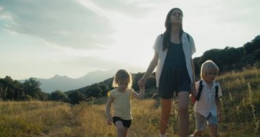 Female traveler holding children hand on summer vacation trip. Family hiking together on travel adventure. Young woman walk on meadow with backpack and kids. Sun flare. 