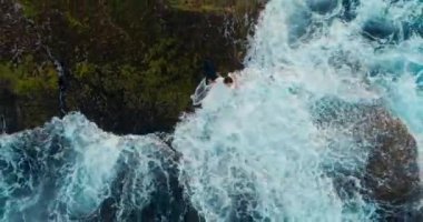 Aerial shot young adult woman in white dress sitting on cliff at the sea shore washed by the waves. Drone flies upwards top to down angle. Concept of summer vacation on coast