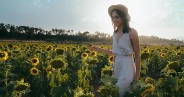 Happy romantic lady walk on sunflower meadow touching flowers and smile on summer vacation. Joyful female tourist in dress walk through plants on journey. Young adult woman with hat on sunny day
