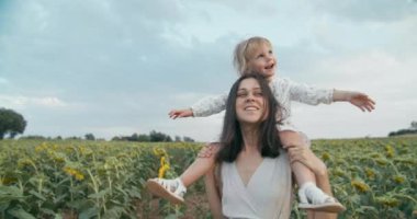 Cheerful young adult woman holding her daughter on neck and spinning around. Family dancing on summer vacation in sunflower meadow surrounded by nature. Joyful children on mother neck