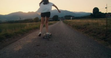 Camera following trendy girl riding on skate longboard at sunset on beautiful mountain view in countryside. Happy young stylish woman skateboarding at evening on summer vacation