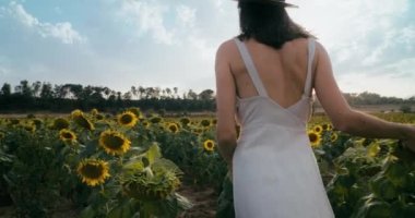 Romantic beautiful lady on summer vacation travel walk on sunflowers meadow. Slow motion camera following young adult woman from back. Feeling free and relaxed girl in hat