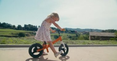 Happy child riding balance bicycle along the agricultural meadow on rural environment. Toddler girl in dress drive bike on sunny summer day. Carefree childhood on countryside. 