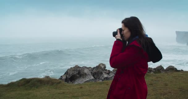 女性旅行者使用模拟摄影设备拍摄美丽的风景 在暴风雨的一天 穿着红色夹克的女人在去海洋的路上给大浪拍照 — 图库视频影像