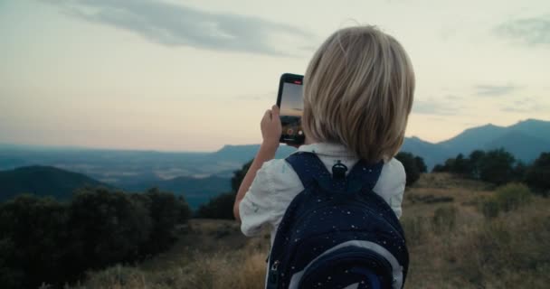 スマートフォンを使用して日没時に山の上のバックパックで休暇旅行中の子供たち 休暇旅行の冒険の間にスマートフォンで撮影した子供の男の子 — ストック動画