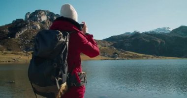 Bahar tatili gezisindeki kadın gezgin kırmızı ceketli göl kıyısında duruyor. Sırt çantasıyla kamerada güzel dağ manzarası fotoğrafı çekiyor. Neşeli kaygısız kadın hafta sonu macerasına çıkıyor.