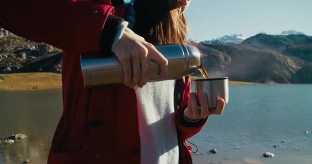 紅色のジャケットと白い帽子の女性は マウンテンレイクショアに立って熱から熱い飲み物を注ぎ ハイキングの旅でそれを飲みます 観光の途中で休憩を取り 自由を楽しむ女性旅行者 — ストック動画