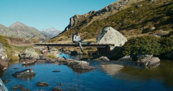 Mężczyzna Blogger Podróży Siedzieć Górskim Drewnianym Moście Nad Rzeką Lub — Wideo stockowe