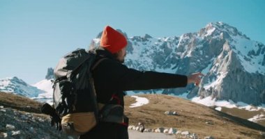 Kış tatilinde sırt çantasıyla gezen genç bir adam. Etrafı görkemli dağ manzarasıyla çevrili. Elleriyle işaret ediyor. Erkek gezgin macerada özgürlüğün tadını çıkarıyor.