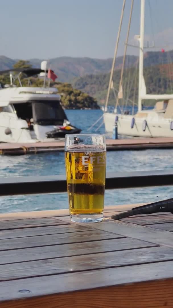土耳其，萨尔萨拉，2021年10月3日：桌上放着一杯啤酒的垂直镜头，美丽的马尔马里斯湾，停泊在海岸线附近的豪华汽艇，斜坡上长着绿树 — 图库视频影像