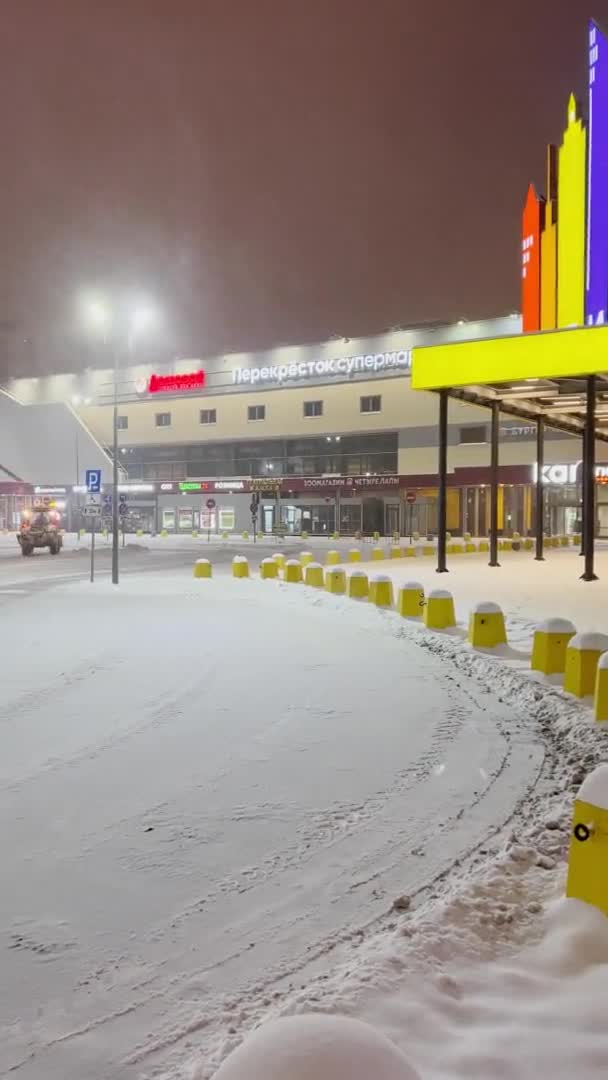 Schneepflug-Traktor beseitigt Schnee von der Fahrbahn auf dem Parkplatz in der Nähe des riesigen Einkaufszentrums, während es nachts keine Autos gibt, nächtliche Beleuchtung, Schneefall, Handelszentrum, Schneesturm — Stockvideo