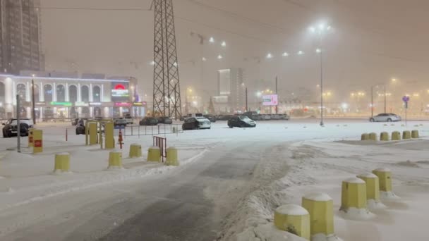 Russland, St. Petersburg, 11. desember 2021: Tungt snøfall i bytrafikken om natten, bilparkering på et stort kjøpesenter, snøstorm, snøstorm, mangefargede skilt på bygningen – stockvideo