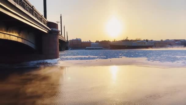 ロシア、サンクトペテルブルク、 2021年12月7日:日没時のサンクトペテルブルクの冬景色のスローモーション映像、凍結したネヴァ川の蒸気、ブラゴヴェシェンスキー橋の近くに係留された巨大な船 — ストック動画