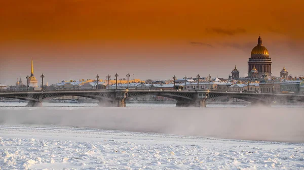 Winter panoramic view of St. Petersburg at sunset, Isaac cathedral and Blagoveshenskiy bridge on background, steam over frozen Neva river, sky of orange color — Fotografia de Stock