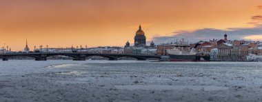 Gün batımında St. Petersburg 'un panoramik manzarası, arka planda Isaac Katedrali ve Blagoveshenskiy köprüsü, donmuş Neva nehrinin üzerinde buhar, setin yanında demirlemiş büyük bir gemi, turuncu renkli gökyüzü.