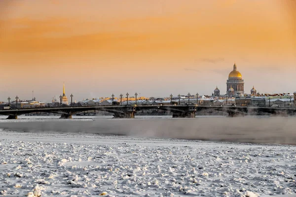 Gün batımında St. Petersburg 'un panoramik manzarası, arka planda Isaac Katedrali ve Blagoveshenskiy köprüsü, donmuş Neva nehri üzerinde buhar, turuncu renkli gökyüzü